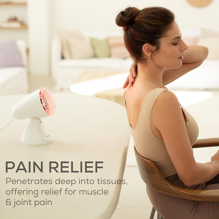 A woman sits in a chair in a bright living room, holding her neck as if relieving muscle pain from the IL11 heat lamp on a table nearby