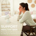 A woman sits at a table in a bright, softly lit room, eyes closed, and facing IL11. IL11 is treating her face to boost skin appearance and support a radiant glow. 